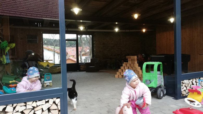 Ein Kind in Winterkleidung spielt mit einem Spielzeugpferd in einem offenen Raum mit Holzstapeln und Spielzeug. Eine Katze läuft vorbei.