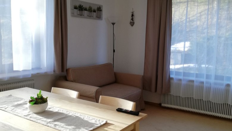 Helles Wohnzimmer mit Esstisch, Stühlen, Sofa und großen Fenstern mit Vorhängen.
