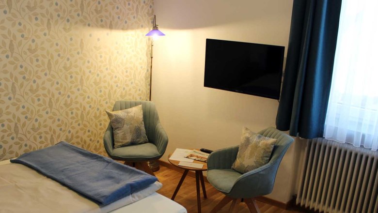 Zimmer mit Bett, Tisch mit zwei Stühlen, Fernseher, Fenster mit blauen Vorhängen und Stehlampe.