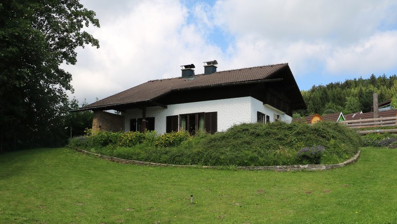 Ein weißes Haus mit braunem Dach in einem grünen Garten mit Bäumen und Blumen.