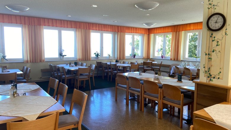 Ein heller Speisesaal mit Holzmöbeln, Tischen und Stühlen, dekoriert mit Pflanzen und einer Wanduhr.