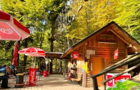 Holzhütte mit Sonnenschirmen und Kletterpark im Hintergrund.