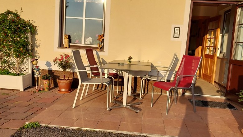 Terrasse mit Tisch und St&uuml;hlen vor einem Geb&auml;ude, umgeben von Pflanzen.