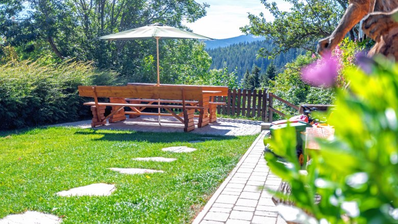 Garten mit Holztisch, Bänken und Sonnenschirm, umgeben von Bäumen und Wiese.