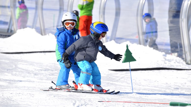 Zwei Kinder in Skiausrüstung fahren im Schnee, begleitet von einem Erwachsenen im Hintergrund.