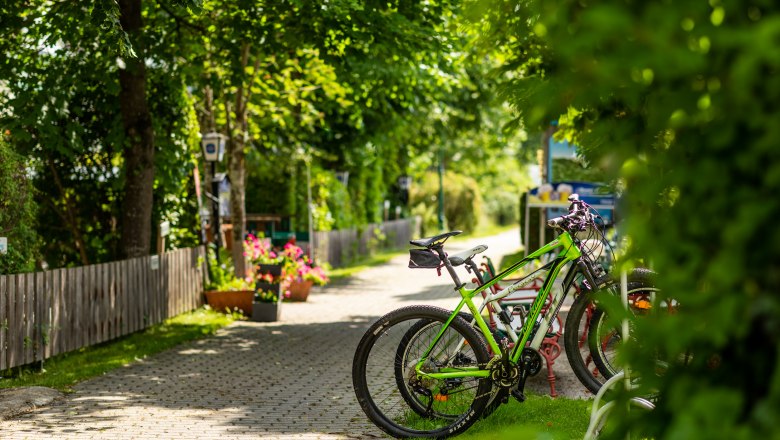 Fahrräder geparkt auf einem Radparkplatz bei einem Gasthof, umgeben von grünen Bäumen und Blumen.