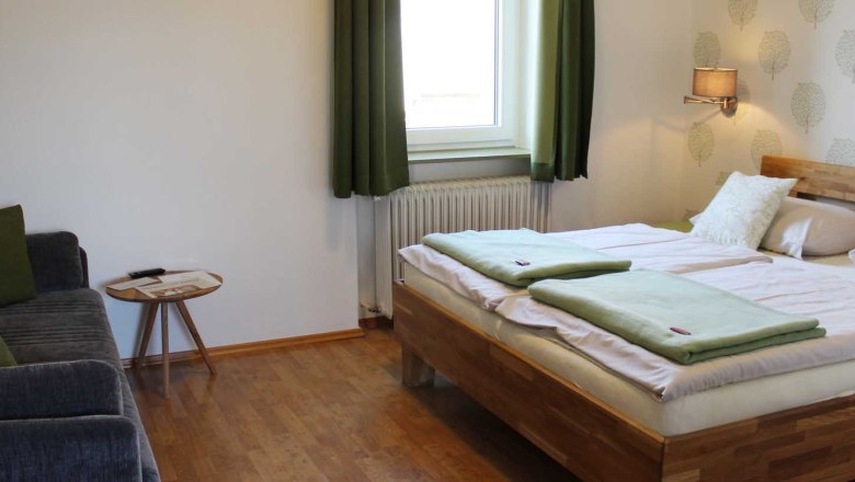 Schlafzimmer mit Doppelbett, Fenster mit gr&uuml;nen Vorh&auml;ngen, Sofa, Tisch, Tapete und einer Wandlampe.