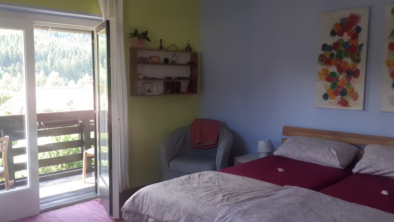 Ein gem&uuml;tliches Doppelzimmer mit zwei Betten, einem Balkon mit Blick auf B&auml;ume und bunte Kunstwerke an der Wand.