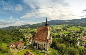Luftaufnahme einer Kirche in einer ländlichen Umgebung mit Hügeln und Wäldern im Hintergrund.