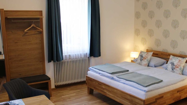 Gemütliches Doppelzimmer mit Bett, Kleiderschrank, Tisch, Stuhl und Fenster mit blauen Vorhängen.