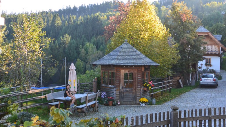 L&auml;ndlicher Hof mit Holzh&uuml;tte, Holzbank, Holzzaun und grauem Auto, im Hintergrund ein Wald.