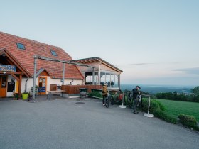 Ein Gasthaus mit Terrasse und Fahrradfahrern in einer l&auml;ndlichen Umgebung.