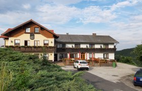 Ein traditionelles Gasthaus mit Balkon und Blumenkästen, davor ein Parkplatz mit Autos, umgeben von grüner Landschaft und blauem Himmel.