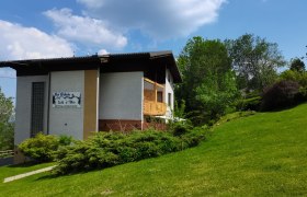 Haus mit Schild 'Bei Webels Ski & Bike' auf gr&uuml;ner Wiese, umgeben von B&auml;umen, unter blauem Himmel.