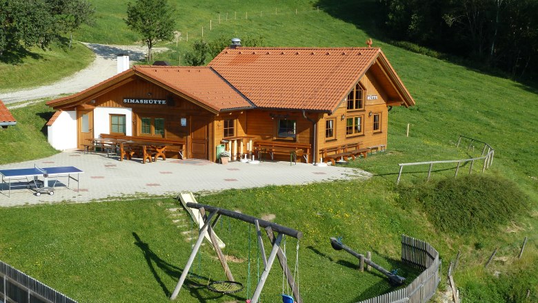 Eine Bergh&uuml;tte namens Simash&uuml;tte mit rotem Dach, umgeben von gr&uuml;nen Wiesen und einem Spielplatz mit Schaukel.