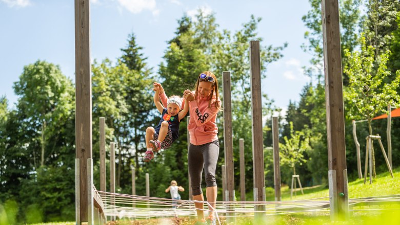 Eine Frau hält ein Kind an den Händen in einem Outdoor-Spielbereich mit Holzpfosten und Seilen.