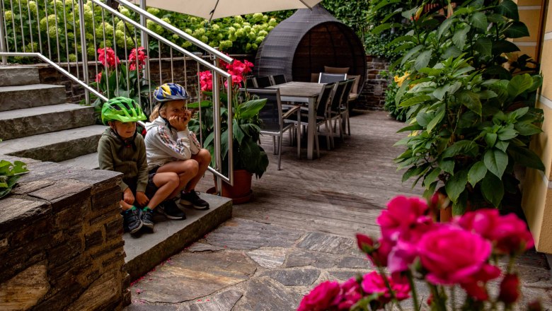 Zwei Kinder mit Fahrradhelmen sitzen auf einer Treppe in einem Garten mit Blumen und Gartenm&ouml;beln.