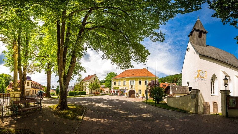 Panoramablick auf den Kirchenplatz von Feistritz mit Kirche, Bäumen und historischen Gebäuden.