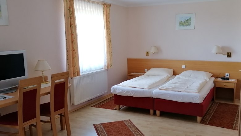 Ein Doppelzimmer mit zwei Einzelbetten, einem Schreibtisch mit Stühlen, einem Fernseher und einem Fenster mit Vorhängen.