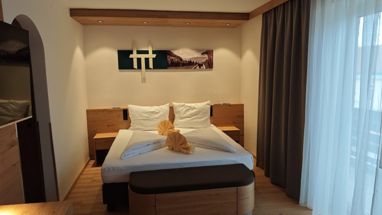 Modern eingerichtetes Doppelzimmer mit einem gro&szlig;en Bett, Holzm&ouml;beln und einem gro&szlig;en Fenster mit Vorh&auml;ngen.