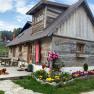 Eine rustikale Holzhütte mit Blumenbeet und einem Hund auf der Terrasse, umgeben von grüner Landschaft und roten Sonnenschirmen.
