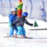 Zwei Kinder in Skiausrüstung fahren im Schnee, begleitet von einem Erwachsenen im Hintergrund.