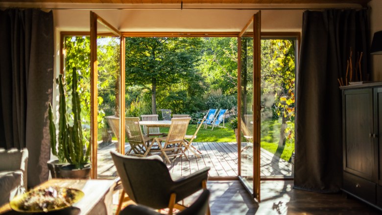 Blick aus einem Wohnzimmer auf eine Terrasse mit Gartenmöbeln und grünem Garten.