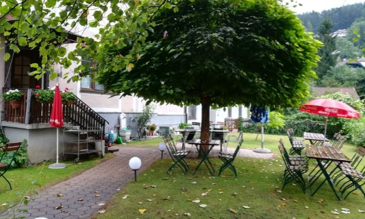 Ein gemütlicher Gastgarten mit Tischen und Stühlen unter einem großen Baum, umgeben von grüner Landschaft.