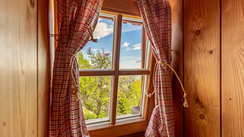 Ausblick aus Holzfenster mit rotem Vorhang auf B&auml;ume unter blauem Himmel.