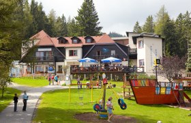 Ein Alpengasthof mit Terrasse und Spielplatz in einer grünen, bewaldeten Umgebung.