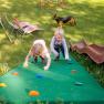 Zwei Kinder klettern auf einem Spielplatz eine grüne Kletterwand hinauf, während ein Hund im Hintergrund steht.