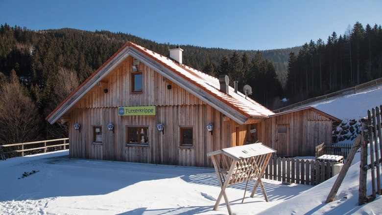 Holzhütte im Schnee mit Schild 'Futterkrippe', umgeben von Wald.