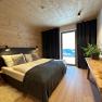 Gemütliches Schlafzimmer mit Holzwänden, Doppelbett, Holzstuhl, Tisch mit Pflanze und großem Fenster mit Vorhängen.