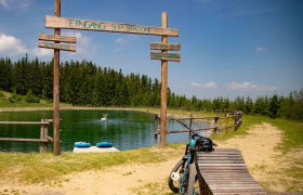 Speicherteich mit SUP-Station in St. Corona am Wechsel, umgeben von Wald und einem Fahrrad im Vordergrund.