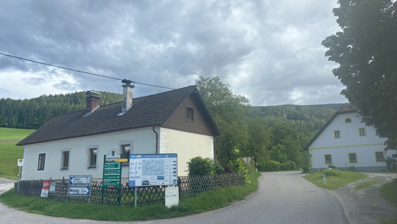 L&auml;ndliche Stra&szlig;e mit zwei H&auml;usern und Wegweisern, umgeben von W&auml;ldern und Wiesen.