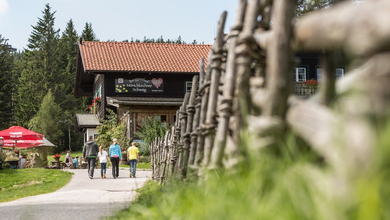 Drei Personen gehen auf einen Berggasthof zu, umgeben von einem Holzzaun und B&auml;umen.