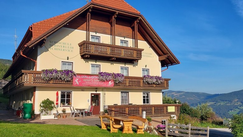 Ein traditionelles Gasthaus mit Holzbalkonen und Blumen, umgeben von gr&uuml;nen H&uuml;geln und blauem Himmel.
