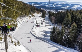 Skigebiet mit Sessellift und Skifahrern, umgeben von schneebedeckten Bäumen und Hügeln.