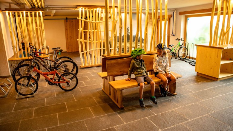 Zwei Kinder mit Fahrradhelmen sitzen auf einer Holzbank in einem Raum mit Fahrr&auml;dern und Holzdekor.
