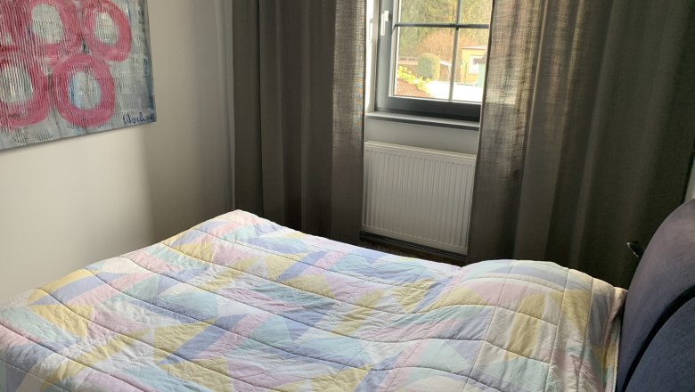 Schlafzimmer mit Bett, bunter Decke, Fenster und Kunstwerk an der Wand.