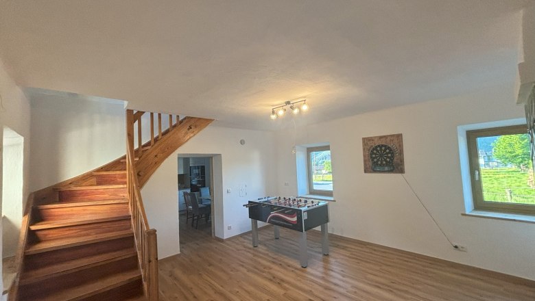 Ein Gemeinschaftsraum mit Tischfu&szlig;balltisch, Dartscheibe an der Wand, Holztreppe und zwei Fenstern.