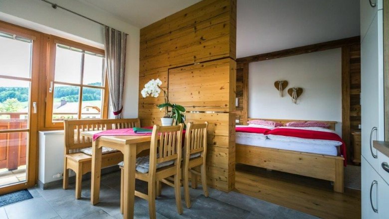 Gem&uuml;tliches Zimmer mit Holzm&ouml;beln, Esstisch, St&uuml;hlen und einem Bett. Fenster mit Blick auf gr&uuml;ne Landschaft.