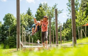 Eine Frau hält ein Kind an den Händen in einem Outdoor-Spielbereich mit Holzpfosten und Seilen.