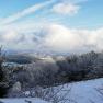 Winterlandschaft mit schneebedeckten Bäumen und Hügeln unter blauem Himmel mit Wolken.
