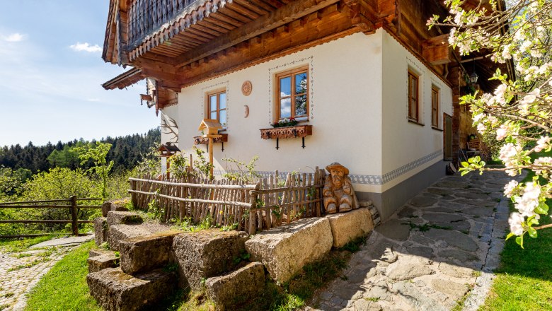 Traditionelles Haus mit Holzbalkon und Gartenzaun, umgeben von Landschaft und bl&uuml;henden B&auml;umen.