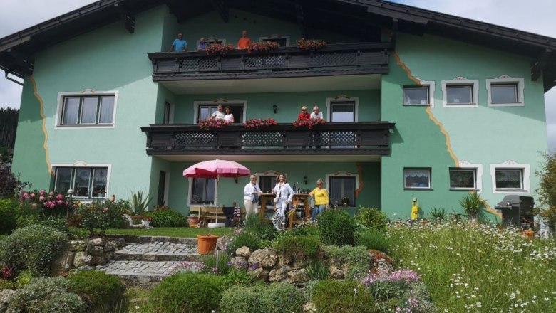 Hausfoto Frühstückspension Buchner Karl, © Buchner Ein grünes Haus mit Balkonen und Menschen davor, umgeben von einem blühenden Garten.