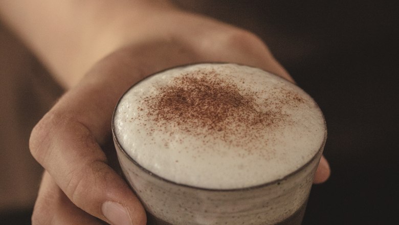 Nahaufnahme einer Hand, die ein Glas mit Cappuccino und Milchschaum h&auml;lt.