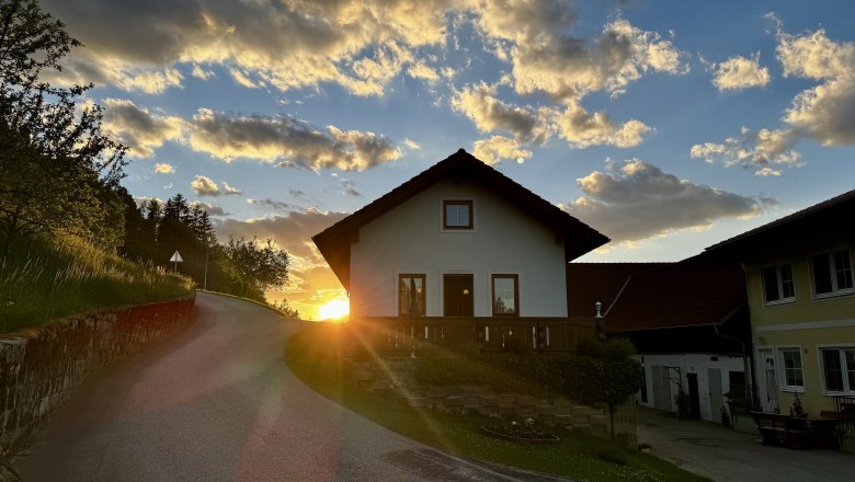 Ferienwohnung Koglhof, © Andrea Tauchner/Koglhof Ein Haus auf einem Hügel bei Sonnenuntergang mit einer kurvigen Straße im Vordergrund.