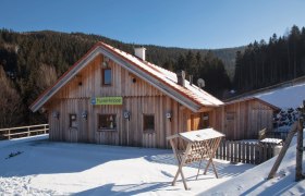 Holzhütte im Schnee mit Schild 'Futterkrippe', umgeben von Wald.