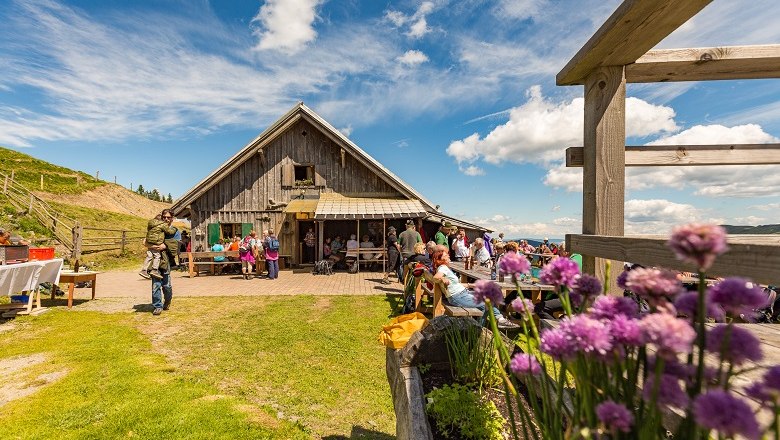 Eine Almhütte mit Menschen, die draußen sitzen, umgeben von Blumen und Wiesen unter einem blauen Himmel.